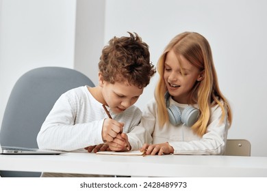 Happy Caucasian children studying at home with their laptops and headphones, immersed in online education The brother and sister are sitting at a table in their cozy living room, focused and connected - Powered by Shutterstock