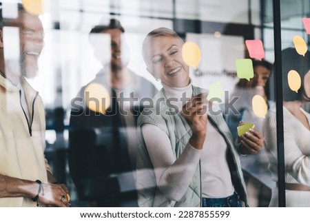 Happy Caucasian colleagues making notes on modern glass board for planning business strategy, collaboration of successful male and female partners during working process in office interior