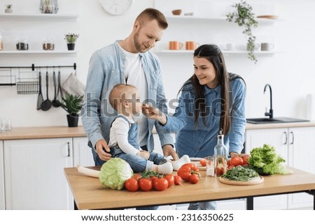 Happy caucasian couple in denim clothes feeding small child with slice of red tomato during lunch time at home. Curious baby girl checking benefits of vegetable rich in vitamin C for proper nutrition.
