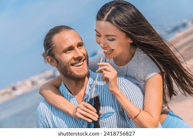 Happy caucasian couple enjoying themselves outdoors. Handsome young guy piggybacking his girlfriend drinking soft drink outdoors by the beach - Powered by Shutterstock