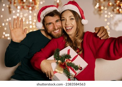 Happy caucasian couple in Santa hats greeting their friends in video call, woman holding gift box, man waving hand to camera - Powered by Shutterstock