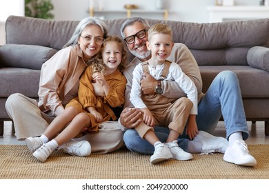 Happy caucasian family grandparents and two cute little kids brother and sister sitting on floor in living room and hugging embracing, looking at camera with smile while spending great time together - Powered by Shutterstock