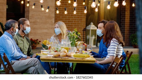 Happy Caucasian Family In Medical Masks Sitting At Table With Meal Outdoor At Picnic And Talking. Joyful Young And Old People Having Dinner And Having Nice Communication. Pandemic Corona Concept.