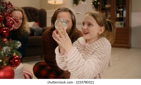 Happy caucasian family opening gifts on Christmas under decorated Christmas tree. Young girl unpacks glass magic snow globe. Warm atmosphere at home on Christmas or New Year. Winter holidays. - Powered by Shutterstock