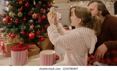 Happy caucasian family opening gifts on Christmas under decorated Christmas tree. Young girl unpacks glass magic snow globe. Warm atmosphere at home on Christmas or New Year. Winter holidays. - Powered by Shutterstock