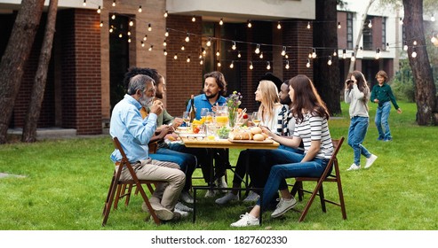 Happy Caucasian family sitting at table with meal outdoor at picnic and talking. Joyful young and old people having dinner and having nice communication. Weekend celebration gathering.