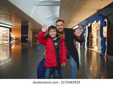 Happy Caucasian Family Of Young Cheerful Dad And Cute Boy Son Waving Looking At Camera While Standing Outside Duty Free Shops In The International Airport Departure Terminal. Family Air Travel Concept