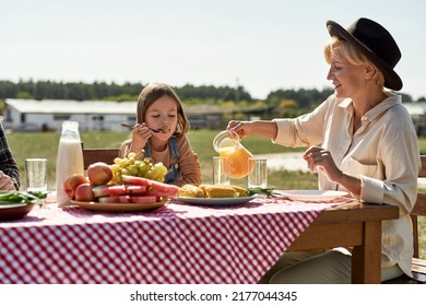 Happy Caucasian Farming Family Having Lunch With Fresh Organic Products In Countryside Outdoors. Grandmother Pouring Juice In Glass Of Granddaughter Who Eating Grape. Modern Farm Lifestyle. Sunny Day