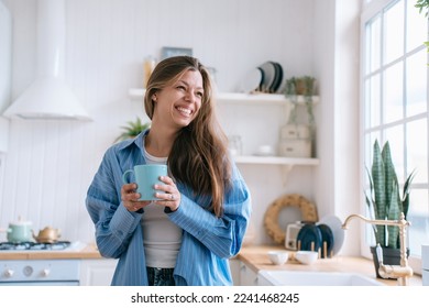 Happy caucasian female with long loose hair in jeans and blue shirt toothy smiles holds cup of tea looks aside happily enjoys weekend at cozy home. Pretty hispanic woman laughs against blurry kitchen. - Powered by Shutterstock