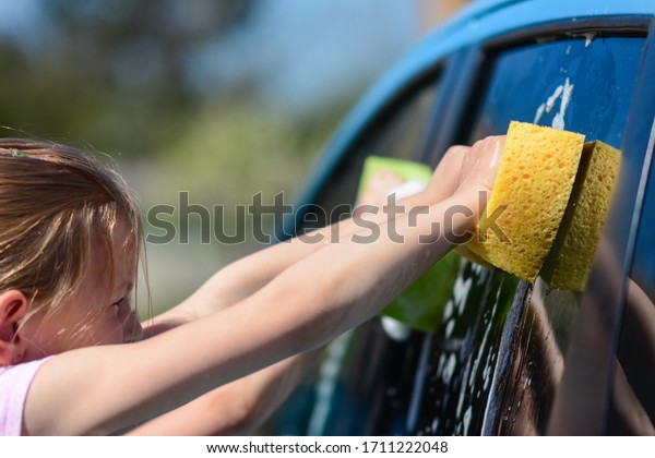 Happy Caucasian girl\
washing car on water splashing and sunlight at home. Elementary\
girl washing a car.