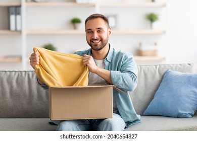 Happy Caucasian Guy Unboxing Cardboard Parcel, Satisfied With Successful Shopping, Taking Out New Sweater, Receiving New Purchase At Home. Online Store And Delivery Concept