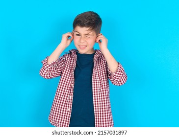 Happy Caucasian Kid Boy Wearing Plaid Shirt Over Blue Background Ignores Loud Music And Plugs Ears With Fingers Asks To Turn Off Sound