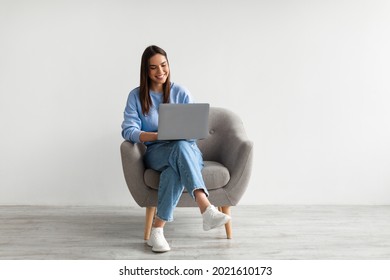 Happy Caucasian lady sitting in armchair with modern laptop, studying remotely or having business meeting against white studio wall, full length. Young woman having online class or watching webinar