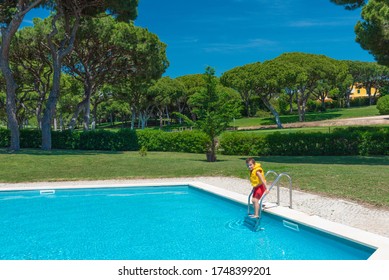 Happy Caucasian Little Boy In Yellow Life Jacket Entering Swimming Pool. Golf Course At The Background. Golf Club With Green Grass. Family Vacation Concept.