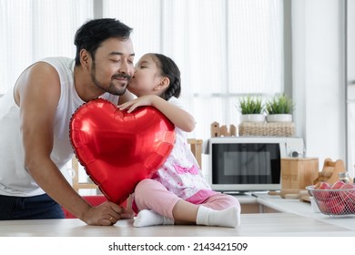 Happy Caucasian little daughter kissing cheek of her Asian young bearded father while holding giving red heart shaped balloon for dad on fathers day at home kitchen with love - Powered by Shutterstock