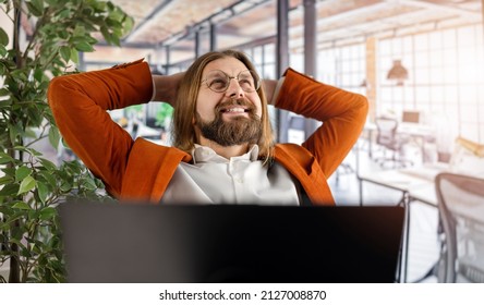 Happy caucasian man in eyeglasses and ornage jacket sitting at desk with modern laptop and keeping hands on head. Bearded male taking break during working process at office.