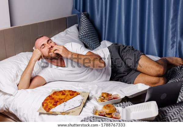 Happy caucasian man having fast food at home in bedroom\
on bed. Man ordered food online take away and eat pizza and burgers\
in comfy room. Hungry bearded tattooed man enjoying fat food.\
