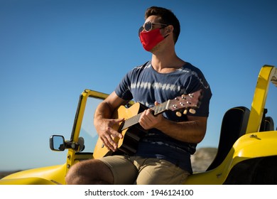 Happy Caucasian Man Wearing Face Mask Sitting On Beach Buggy Playing Guitar. Beach Stop Off On Summer Holiday Road Trip During Coronavirus Covid 19 Pandemic.