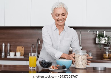 Happy Caucasian mature woman having healthy breakfast at home kitchen. Slim middle-aged housewife preparing oat flakes in the morning with fruits and orange juice - Powered by Shutterstock