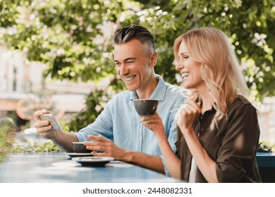 Happy caucasian middle-aged spouses couple hugging embracing bonding and drinking coffee in cafe restaurant while watching social media on cellphone together on a date. Love and relationship concept. - Powered by Shutterstock