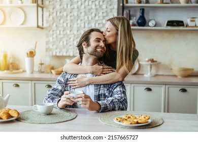 Happy Caucasian Millennial Blonde Wife Kissing Man With Stubble On Light Kitchen Interior, Sun Flare. Couple Enjoy Free Time And Breakfast Together, Family, Relationship, Love And Romance At Home