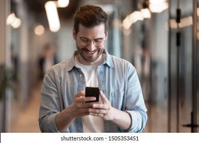 Happy Caucasian Millennial Male Employee In Glasses Stand Hold Cellphone Texting Messaging On Application, Smiling Young Man Worker Using Smartphone Check Email, Browsing Internet Or Web