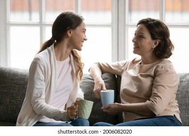 Happy Caucasian mom and adult millennial daughter sit relax on sofa in living room drink tea coffee enjoy family weekend together. Smiling middle-aged mother and grownup girl child rest at home.
