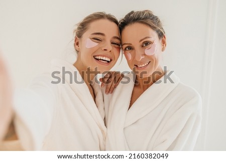 Happy caucasian mother with daughter wearing hydrogel patches smiling looking at camera on white background. Blondes wear robes to take care of their skin. Cosmetic natural trend.