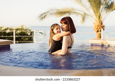 Happy Caucasian Mother And Her Little Cute Daughter Playing In Swimming Pool. Mother And Baby Girl Swim In The Pool. Mom Holding Her Daughter In Her Arms In The Pool And Teaching Her To Swim