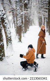 Happy Caucasian Mother Pulling Sledge With Her Cute Little Son While Walking Together Among Snowy Forest. Family Spending Time Together Outdoors During Winter Time.