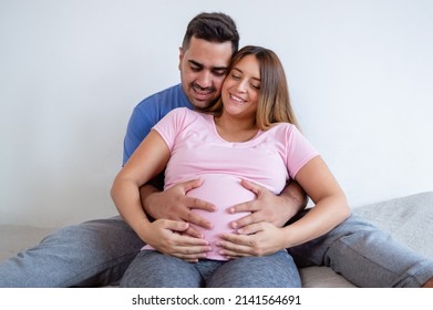 Happy Caucasian Pregnant Woman With Her Husband Sitting On Bed Smiling And Touching Baby On Tummy, Doing Pregnancy Therapy And Exercise Routine.