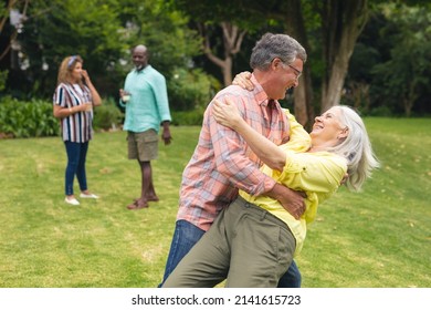 Happy Caucasian Senior Couple Dancing While Friends In Background During Backyard Party. Unaltered, Lifestyle, Leisure, Togetherness, Retirement And Backyard Party.