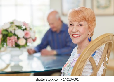 Happy Caucasian Senior Couple In Dining Room. Female Sitting Looking At Camera. Senior Husband Out Of Focus Looking At Papers At Table. Bright And Airy.