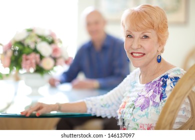 Happy Caucasian Senior Couple In Dining Room. Female Sitting Looking At Camera. Senior Husband Out Of Focus Looking At Camera. Bright And Airy.