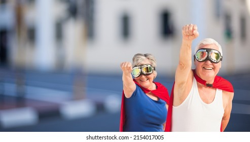 Happy Caucasian Senior Couple Wearing Red Capes And Masks With Hand Raised Standing In City. Love, Togetherness, Superhero, Power, Fantasy, Unaltered, Senior Citizens, Retirement And Awareness.