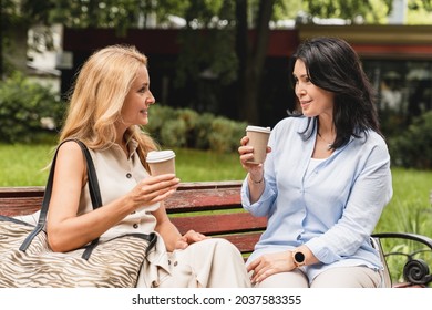 Happy Caucasian Sisters Two Mature Women Girlfriends Best Friends Talking Spending Time Together While Sitting On The Park Bench Drinking Coffee.