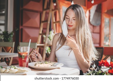 Happy Caucasian Tourist Girl Sits Alone At A Table In A Cafe, Takes Off Her Protective Medical Face Mask And Look At Food, Coronavirus Quarantine Social Distance