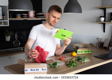 Happy Caucasian White Man Unpack Online Home Food Delivery. Box With Packed Tuna, Shrimp, Vegetables And Recipe Card On A Kitchen Background. Food Delivery Services. Dinner Set From Restaurant