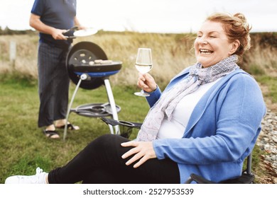 Happy Caucasian woman enjoying a glass of wine outdoors. Woman smiling, holding wine, and relaxing. Man grilling in the background. Woman and wine, outdoor relaxation. Barbecue on camping family trip. - Powered by Shutterstock