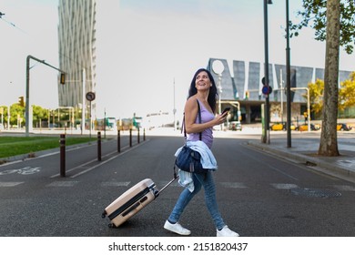 Happy Caucasian Woman Looking Away While Pulling Rolling Suitcase And Using Mobile Phone On City Street