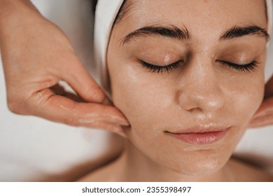 Happy caucasian woman receiving soothing mask or moisturizing cream balm, massaging face at spa salon. Daily spa morning routine.