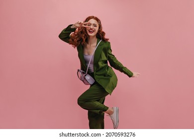 Happy Caucasian Woman Showing Victory Sign Near Face Smiling Strongly On Pink Background. Long-haired Red-haired Beauty Laughs With Her Leg Curled Up At Knee, Dressed In Jacket, Pants And Top.