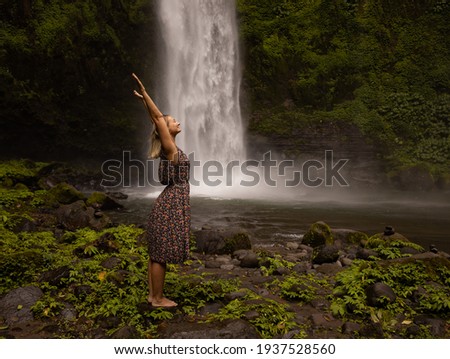 Happy Caucasian woman spending time near waterfall, raising hands up. Nature concept. Energy of water. Travel lifestyle. Woman wearing dress. Copy space. Nung Nung waterfall in Bali, Indonesia