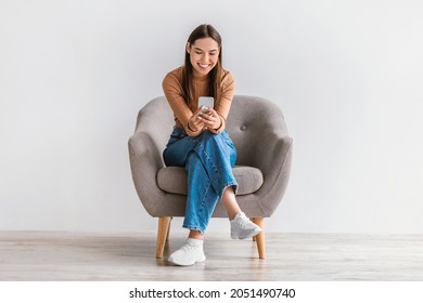 Feliz mujer caucásica usando el celular, chateando en internet, trabajando o estudiando en línea, sentada en sillón contra pared de estudio blanco, de largo. Chica joven vigila video en el teléfono móvil