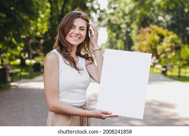 Happy caucasian young brunette woman holding a poster in her hands and smiling on the background of the park - Powered by Shutterstock