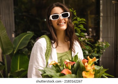 Happy caucasian young girl with snow-white smile looks at camera through sunglasses. Brunette holds flowers in her hands standing outdoors. Rest time concept