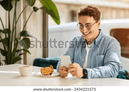 Happy caucasian young man student with curly hair and glasses using his smartphone at a cafe table with a fresh croissant and coffee, enjoying a bright morning indoors. Devise for work