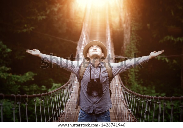 Happy celebrating winning success male tourist\
standing elated with arms raised up above on pedestrian hanging\
bridge over river in tropical\
forest