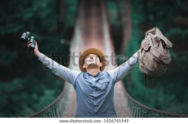 Happy\
celebrating winning success male tourist holding binoculars and\
backpack standing elated with arms raised up above on pedestrian\
hanging bridge over river in tropical\
forest