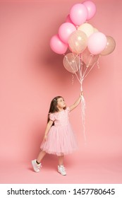 Happy celebration of birthday party with flying balloons of charming cute little girl in tulle dress smiling to camera isolated on pink background. Charming smile, expressing happiness. Place for text